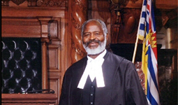 Emery Barnes standing in formal robes in the BC Legislature chamber, with the BC provincial flag in the background.