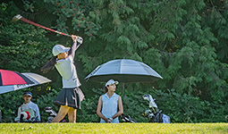 Female golfer executing a powerful swing on a sunny course, observed by onlookers and a caddy, with lush greenery in the background.