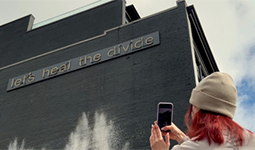 A person with pink hair, wearing a toque, uses their phone to take a photo of fluorescent tubing on the exterior of a black building outlining the words Let's Heal the Divide