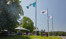MST flag raised on flagpoles during a community gathering with attendees seated under tents in a park.