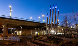 NEU building under the Cambie Bridge at dusk, with illuminated exhaust stacks and raised garden beds in the foreground.