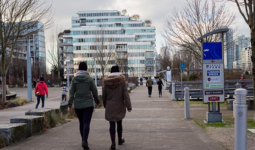 People walking along a paved pathway in Olympic Village, lined with trees and signage. 
