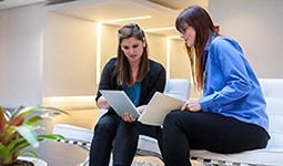 Two women in conversation, seated on office armchairs, both looking at a tablet. One woman holds paperwork.