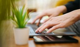 Close-up of hands typing on a laptop keyboard, placed on a desk with a smartphone and a small potted plant.