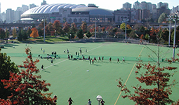 Outdoor synthetic sports field with players and city skyline in the background.