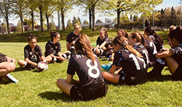 Youth soccer team in black uniforms sitting in a circle on grass, listening to a coach during a sunny day.